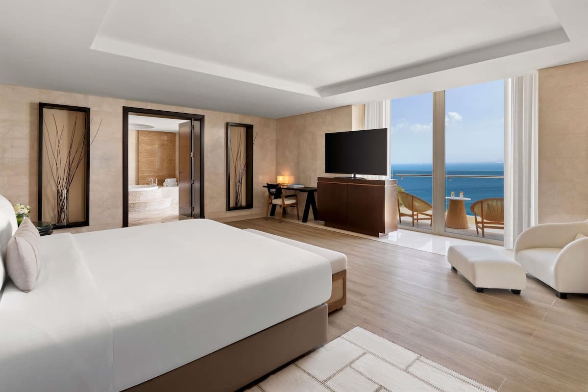 A modern hotel room with a large bed, a TV, a desk, and a balcony with a view of the ocean.