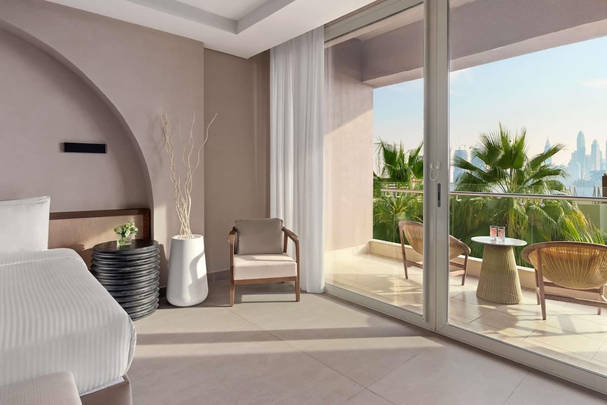 A modern hotel room with a balcony, a bed, a seating area, and a view of the city skyline.