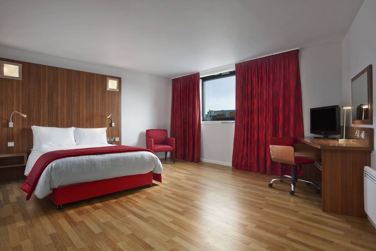 A modern hotel room with a wooden floor, a large bed with red bedding, a desk with a computer, a chair, and a TV.
