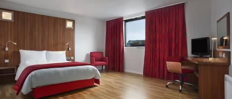 A modern hotel room with a wooden floor, a large bed with red bedding, a desk with a computer, a chair, and a TV.