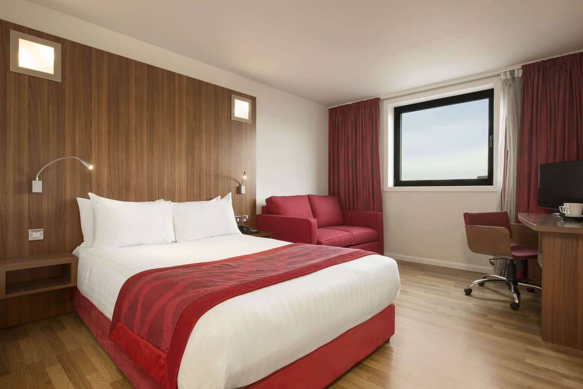 A hotel room with a large bed, a desk, a chair, a TV, and a window with curtains.