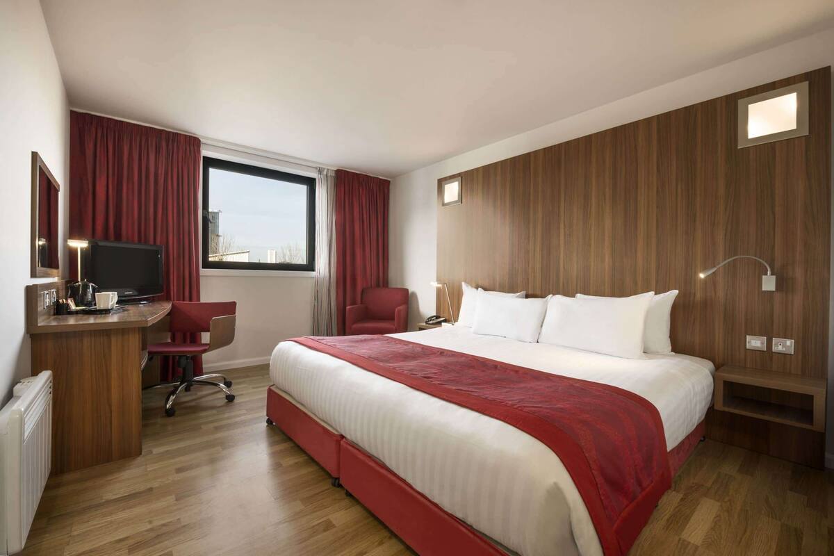 A hotel room with a large bed, a desk with a TV, a chair, and a window with curtains.