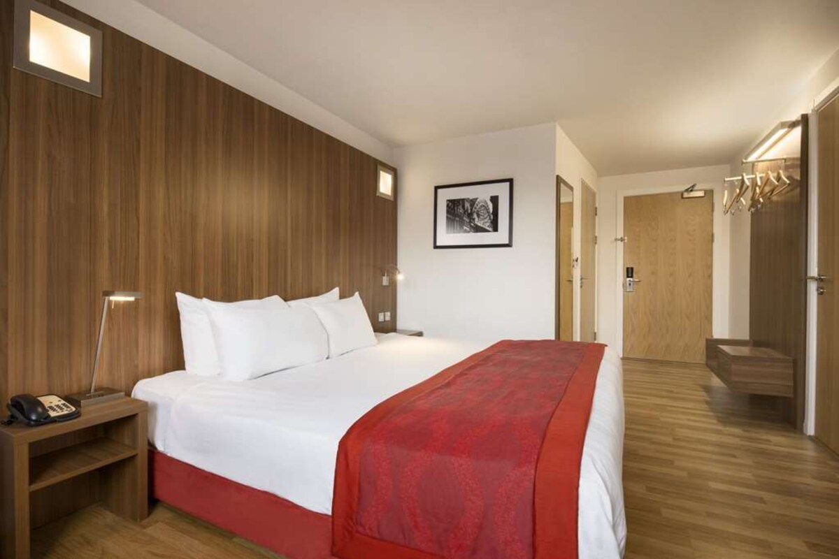 Een hotelkamer met een groot bed, een nachtkastje met een telefoon, een rode plaid en een ingelijst schilderij aan de muur.