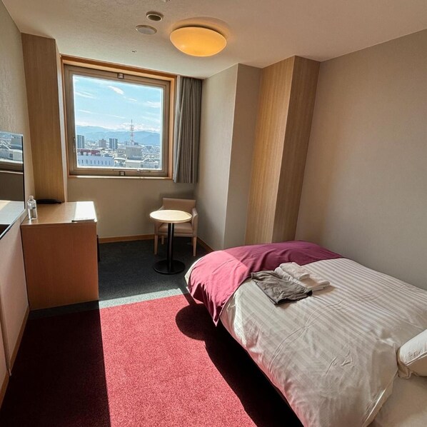 A hotel room with a bed, a desk, a chair, a window with a city view, and a red carpet.