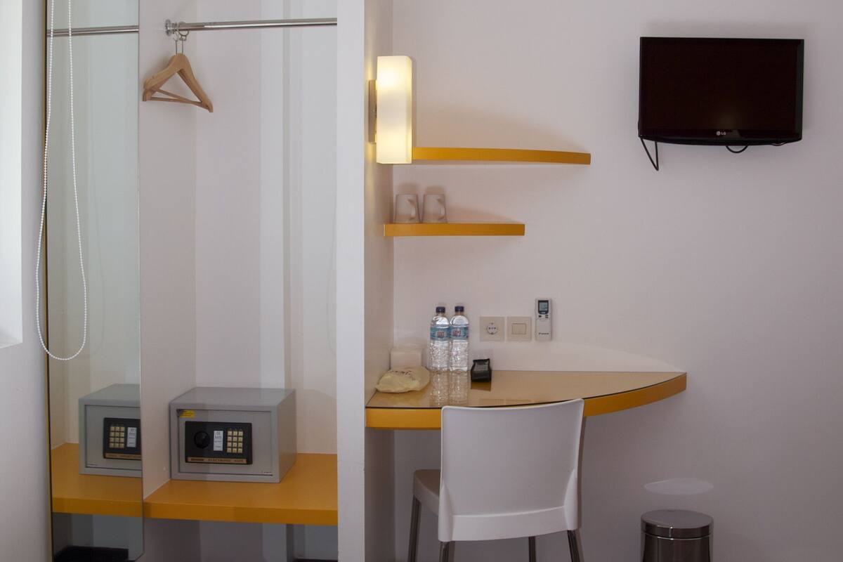 A hotel room with a bed, a desk with a chair, a TV mounted on the wall, a mirror, a hanging shower curtain, a small trash bin, and a heater on the floor.