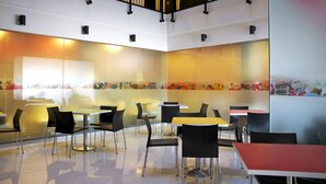 A modern indoor dining area with multiple tables and chairs, a glass wall with colorful artwork, and a staircase with a red railing. - Amaris Hotel Ambon (Ambon)