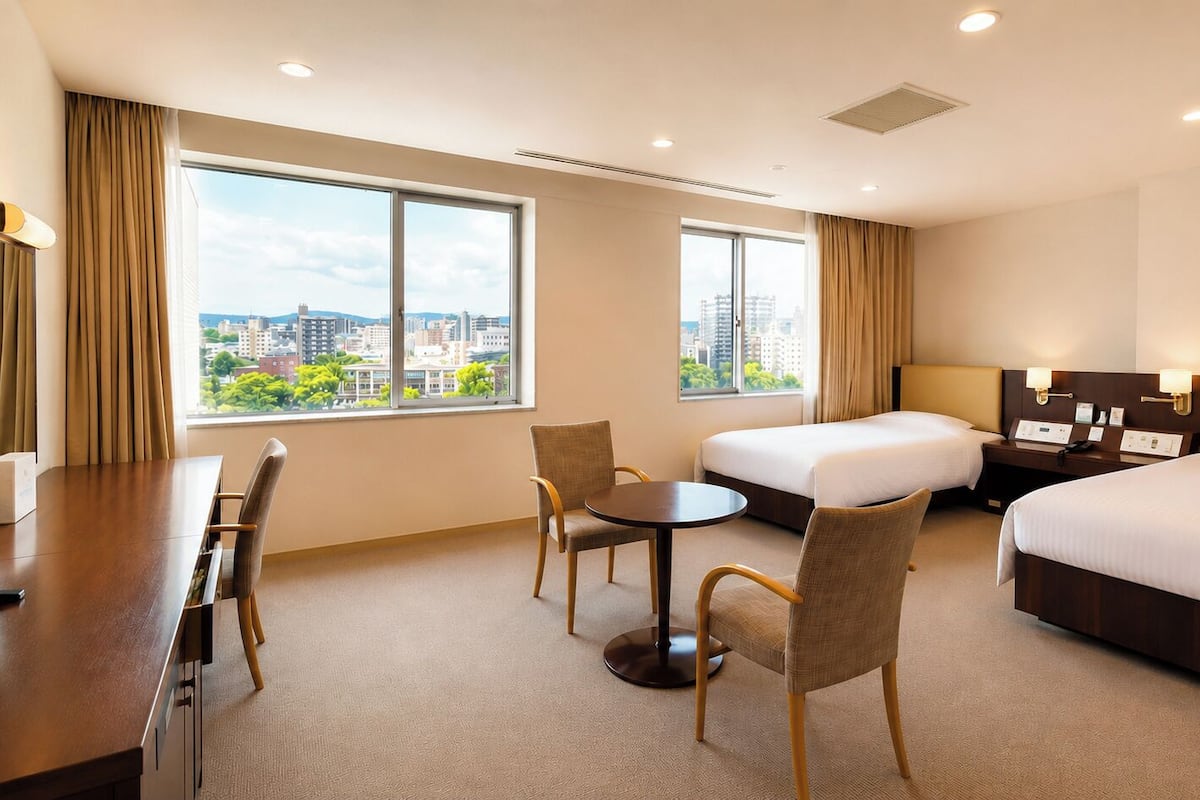 A hotel room with a bed, a desk with a TV, a small table with chairs, and large windows with a city view.