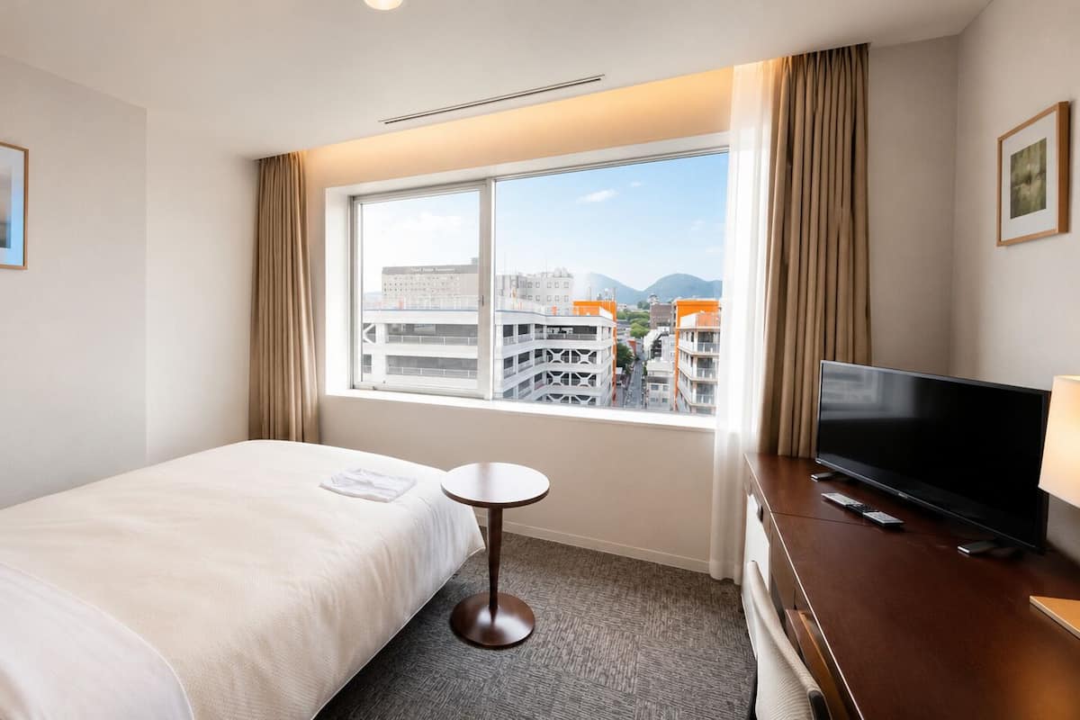 Een hotelkamer met een bed, een klein tafeltje, een televisie en een raam met uitzicht op de stad.
