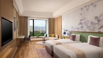 A hotel room with two beds, a TV, a desk with a chair, and a large window offering a city view.