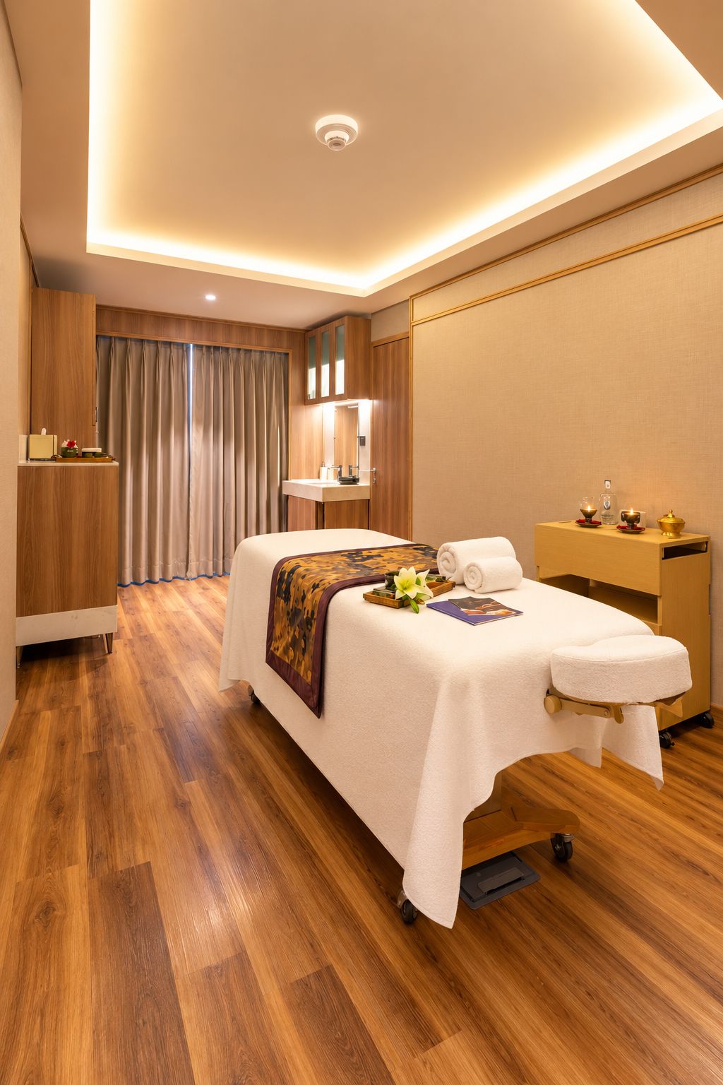 Habitación de hotel con una camilla de masaje, suelo de madera, tocador con espejo y escritorio con lámpara.