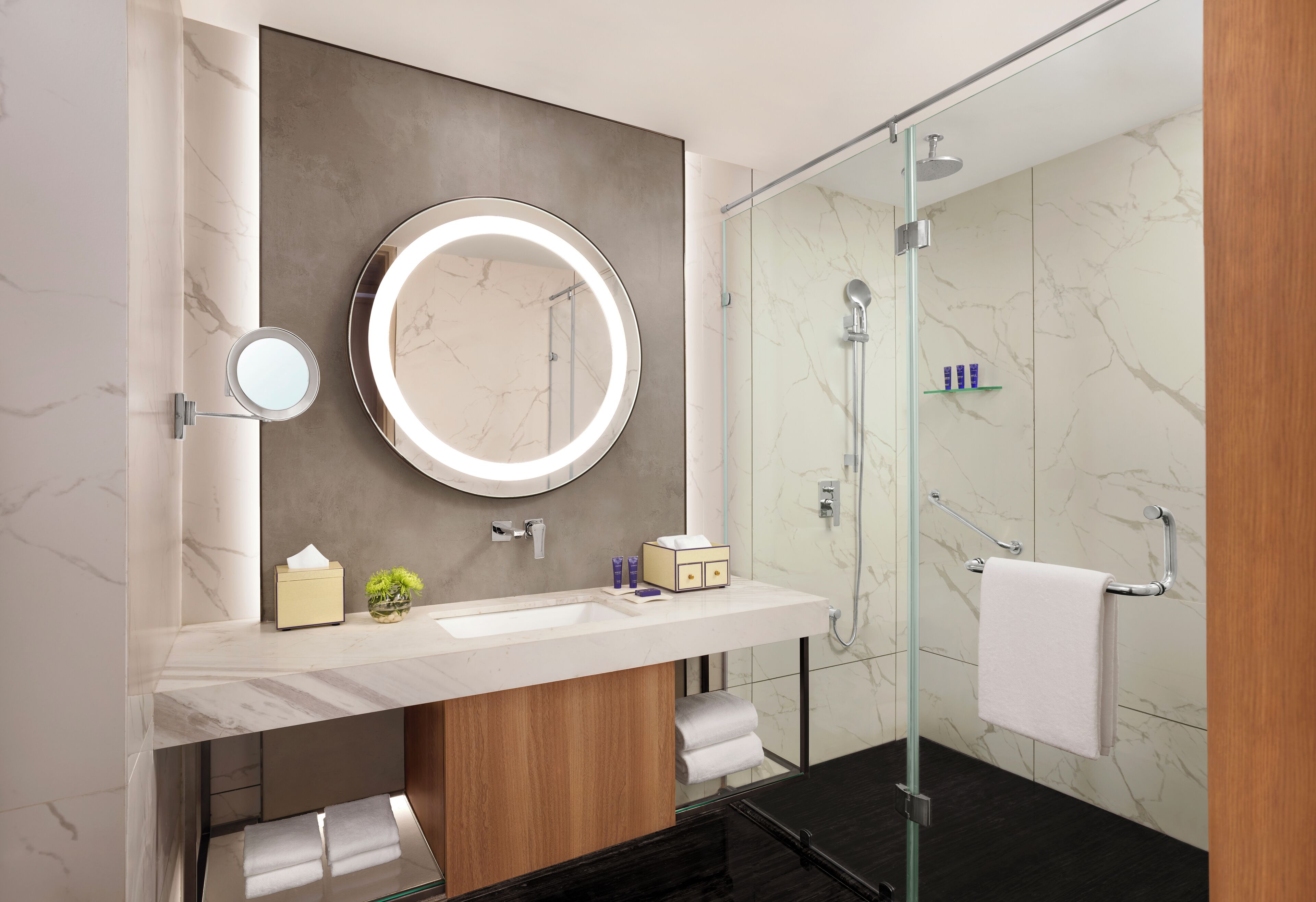 A modern bathroom with a marble vanity, a round mirror with a light, a glass-enclosed shower, and a towel rack.