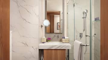 A modern bathroom with a marble wall, a wooden vanity, a round mirror, a glass shower enclosure, and a towel hanging on the door.