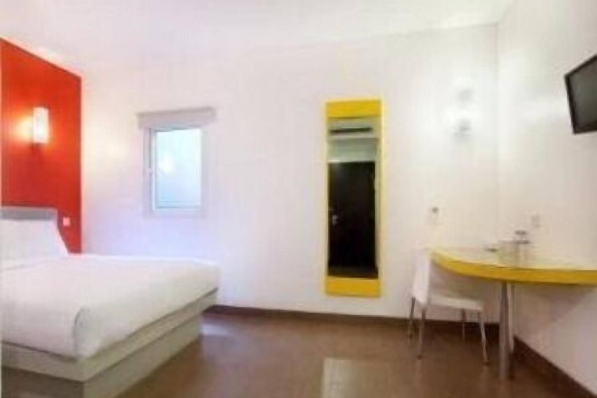 A hotel room with a bed, a desk, a chair, a small window, and a TV mounted on the wall.