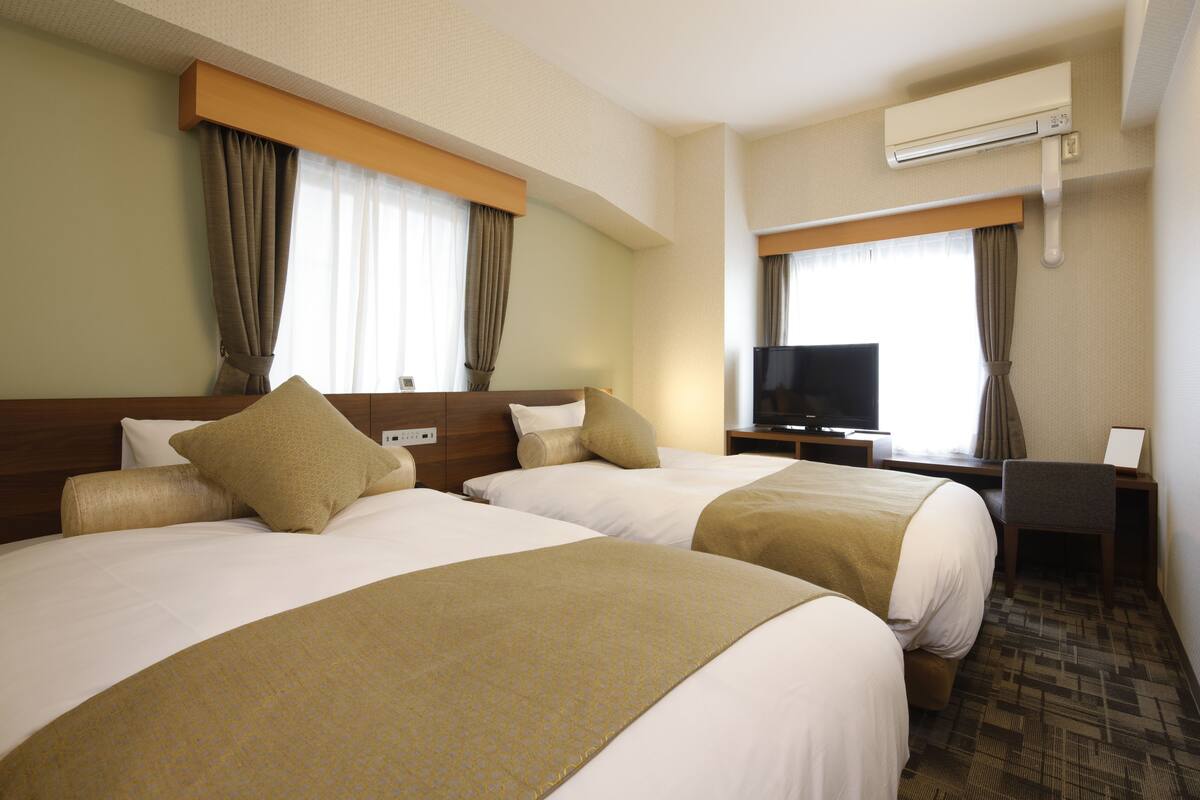 A hotel room with two beds, a television, a desk, and an air conditioning unit.