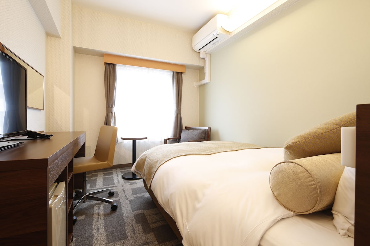 A hotel room with a bed, a desk, a chair, a small table, and an air conditioning unit.