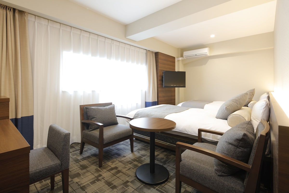A hotel room with a bed, two armchairs, a small table, a TV, and an air conditioning unit.