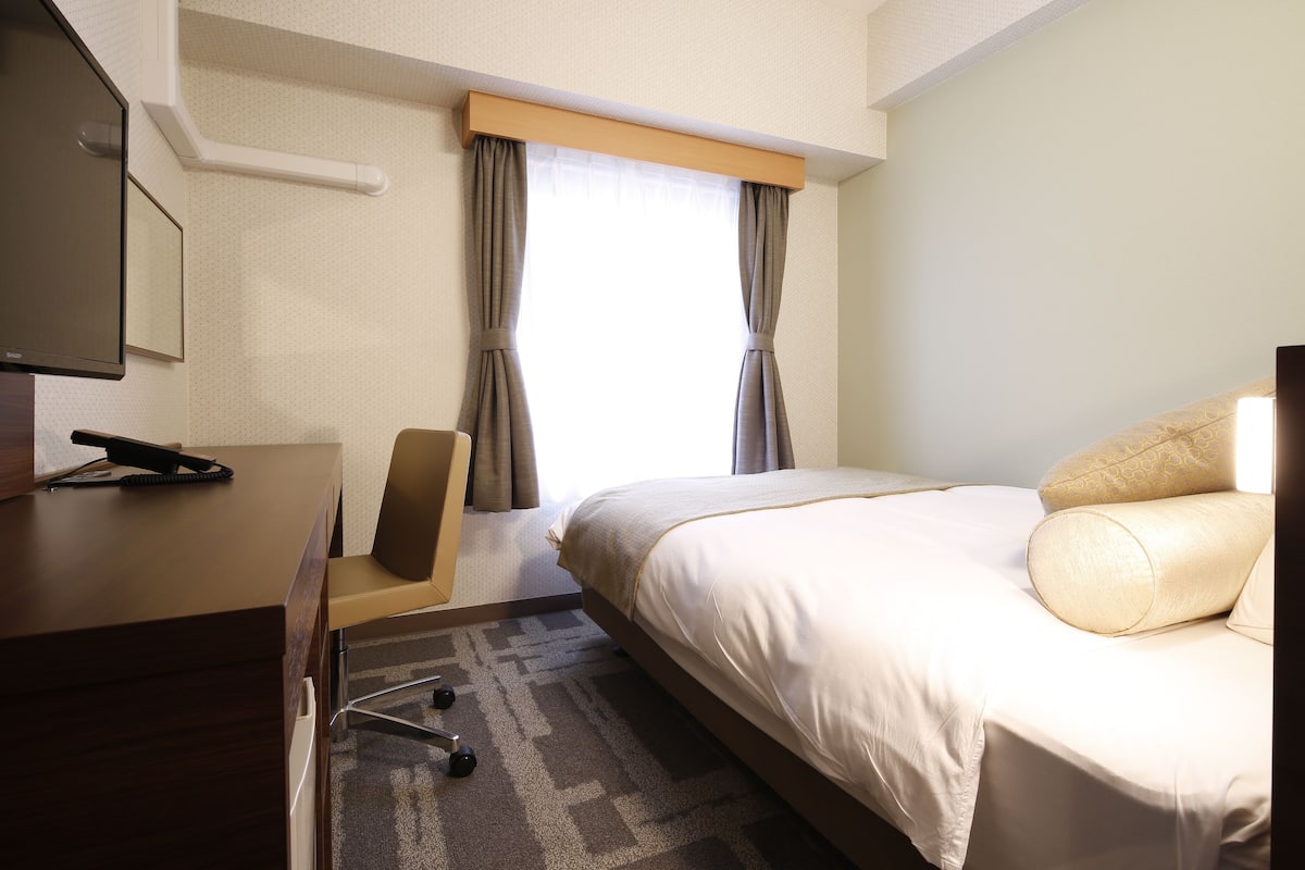 A hotel room with a bed, a desk with a telephone, a chair, and a window with curtains.