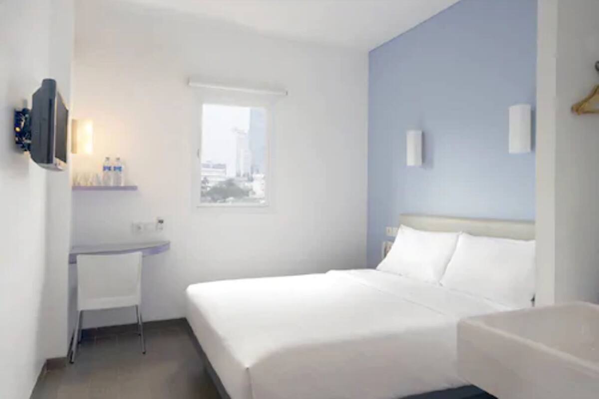 Habitación de hotel con cama, escritorio, silla, ventana con vista a la ciudad y televisor montado en la pared.
