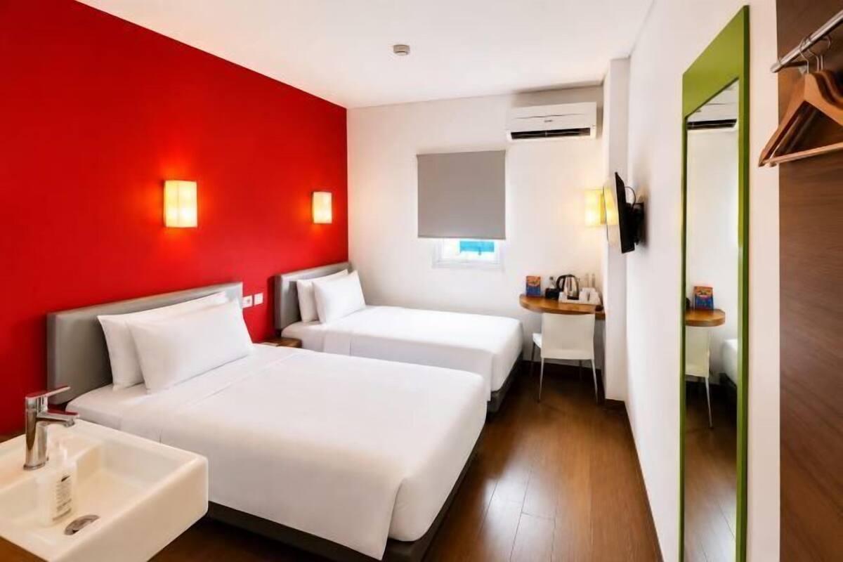 Habitación de hotel con dos camas, un lavamanos, un espejo, un televisor y una pequeña mesa con una lámpara.