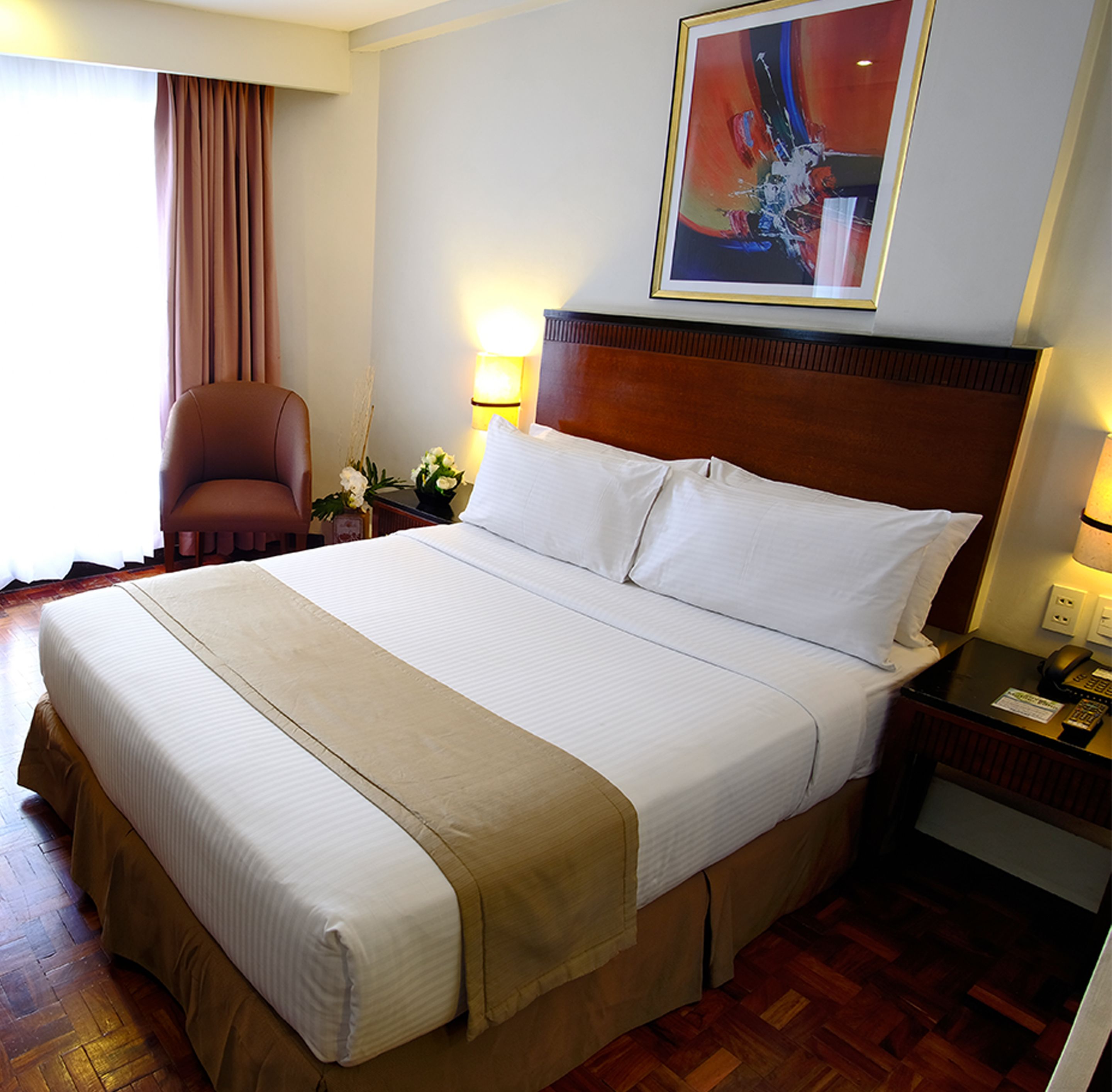 A hotel room with a large bed, a wooden headboard, a nightstand with a phone, a chair, and a painting on the wall.