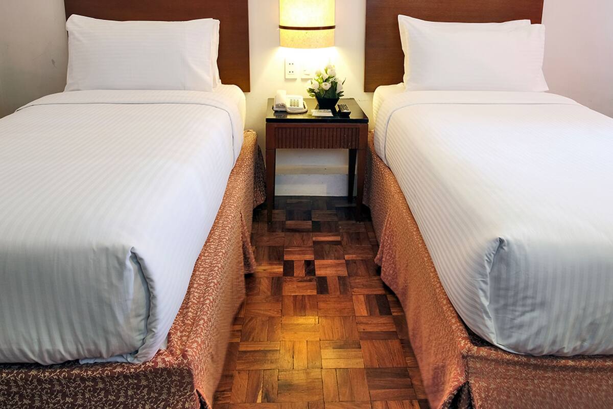 Habitación de hotel con dos camas individuales, cabecera de madera, mesita de noche con lámpara y dos cuadros enmarcados en la pared.