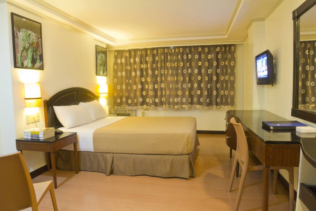 Habitación de hotel con cama, escritorio, silla, televisor y cortinas.