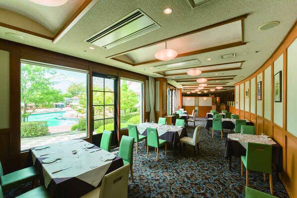 Un comedor de hotel con mesas dispuestas para los huéspedes, amplias ventanas que ofrecen vistas a una piscina y follaje, y una alfombra estampada.
