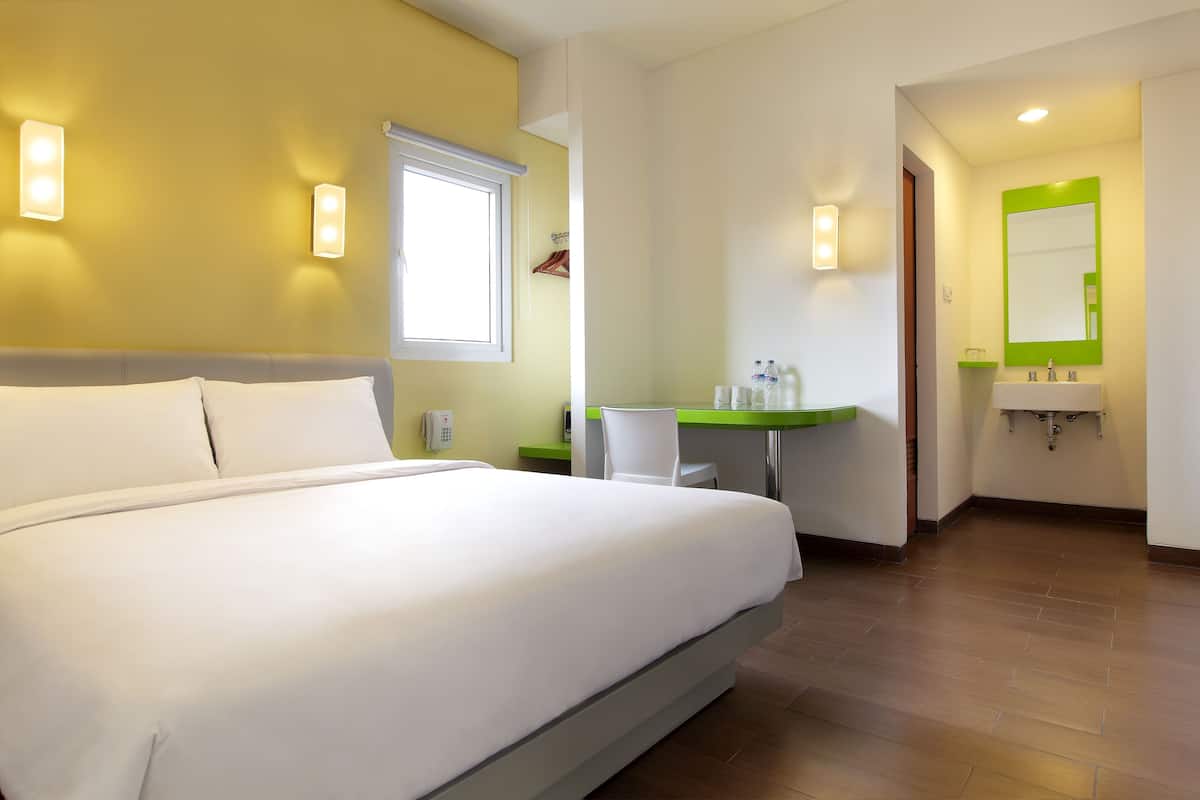 A hotel room with a large bed, a desk with a chair, a bathroom with a sink and mirror, and a window with blinds.