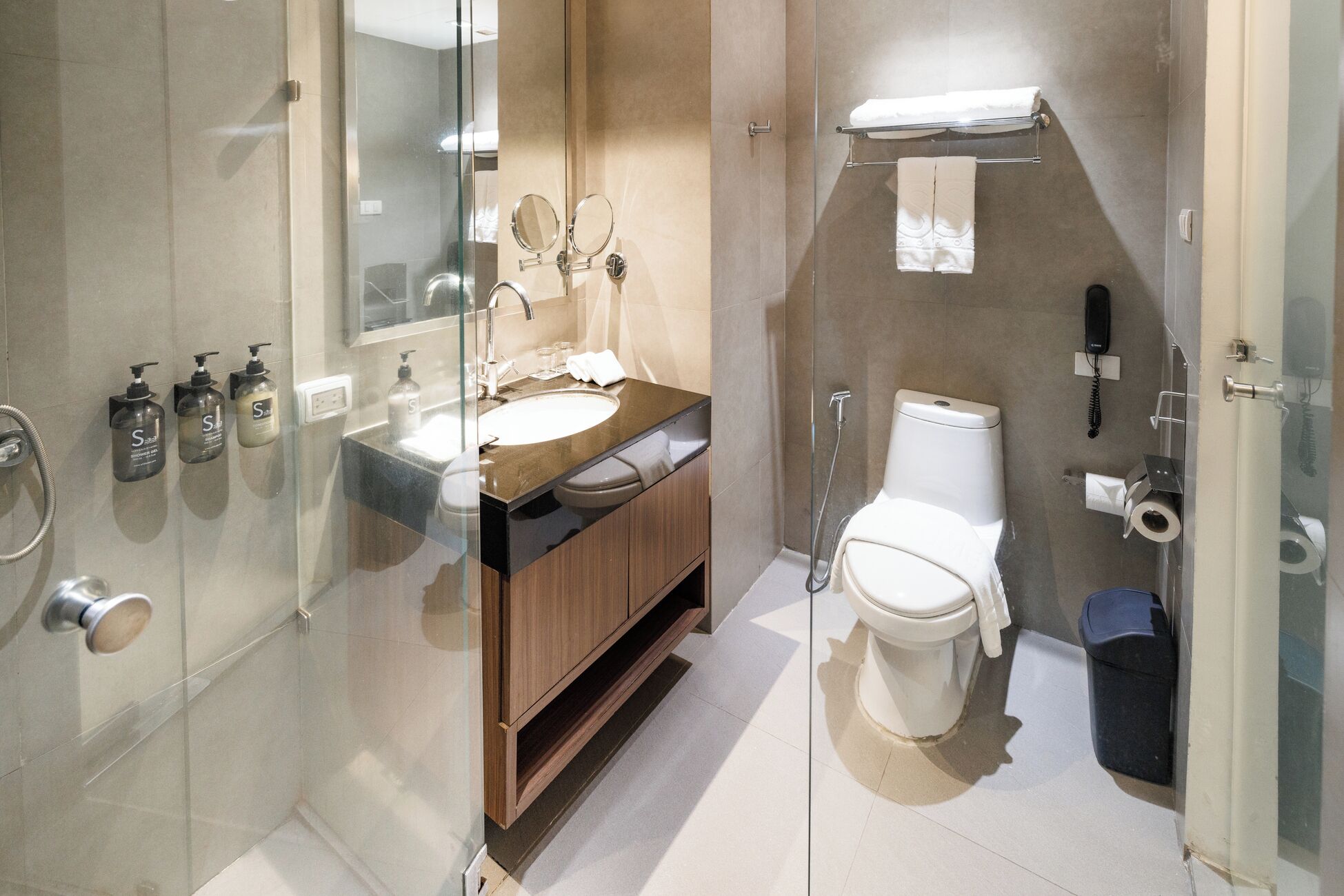 Un baño moderno con cabina de ducha de vidrio, tocador de madera, inodoro blanco y separador de vidrio.
