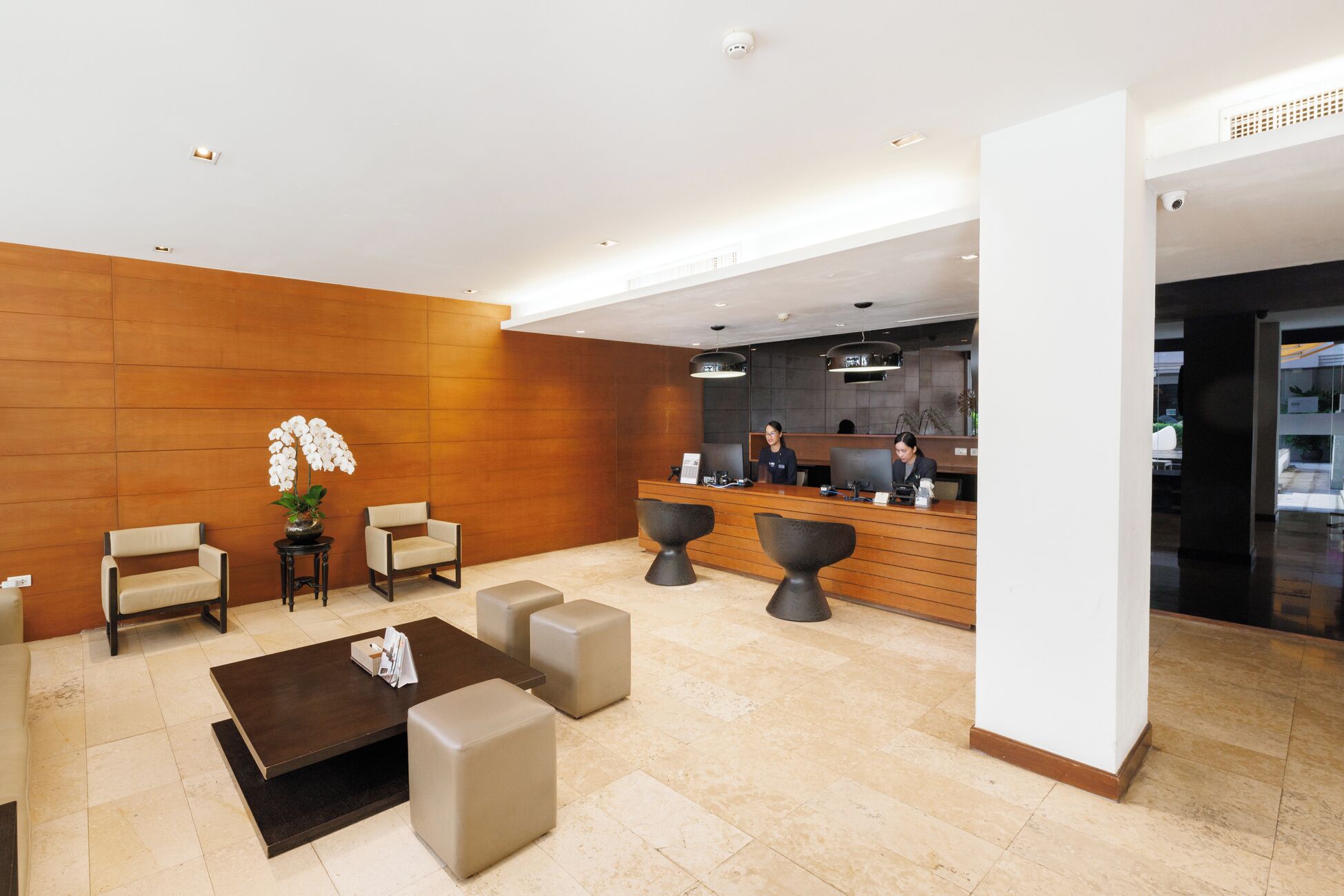 Un área de recepción moderna con un muro de madera, dos recepcionistas, un mostrador de recepción y una zona de estar con una mesa de centro y otomanas.