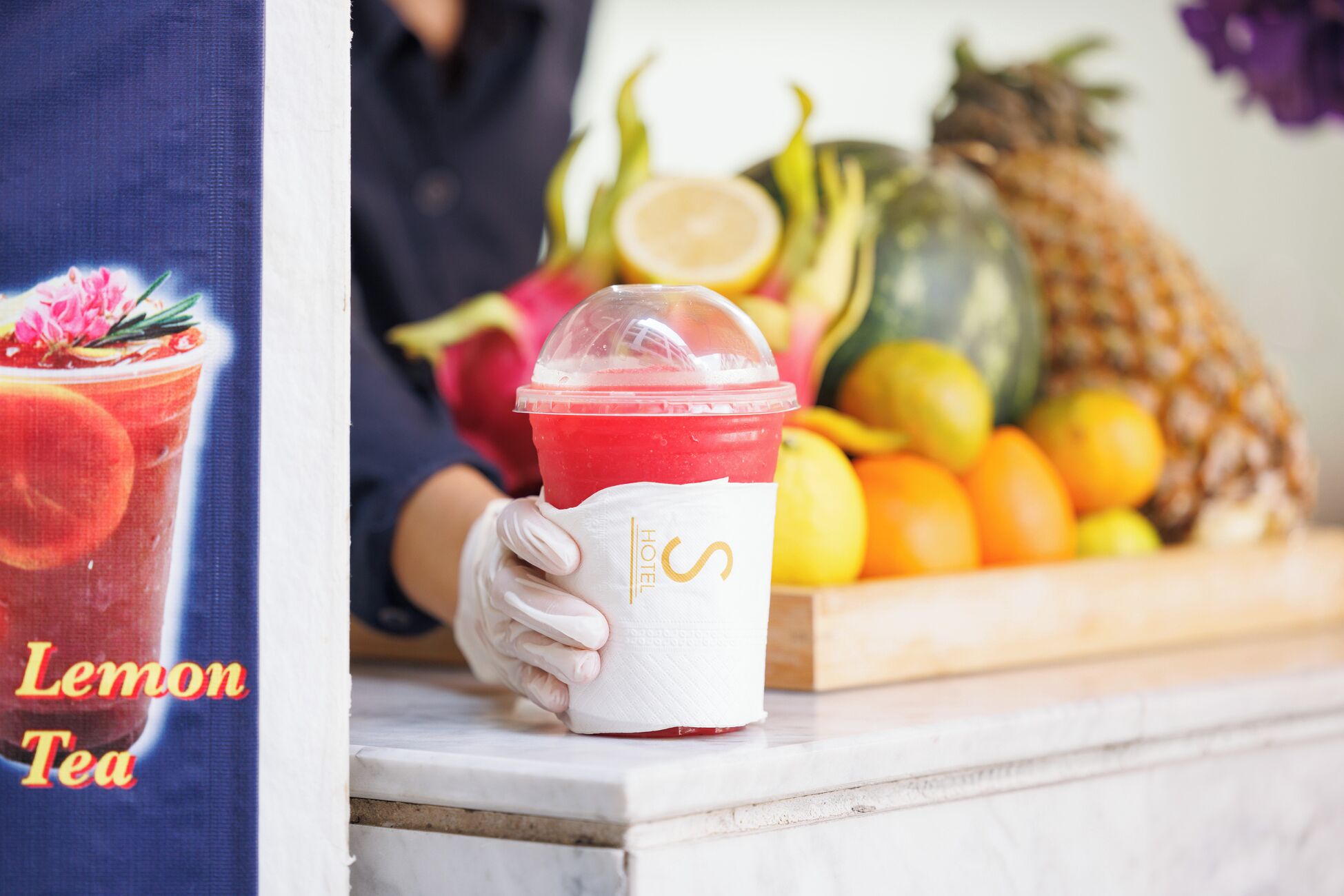 Iemand houdt een rood drankje met een citroenpartje vast, een fruitschaal met verschillende soorten fruit, en een bordje met de tekst 'Lemon Tea'.