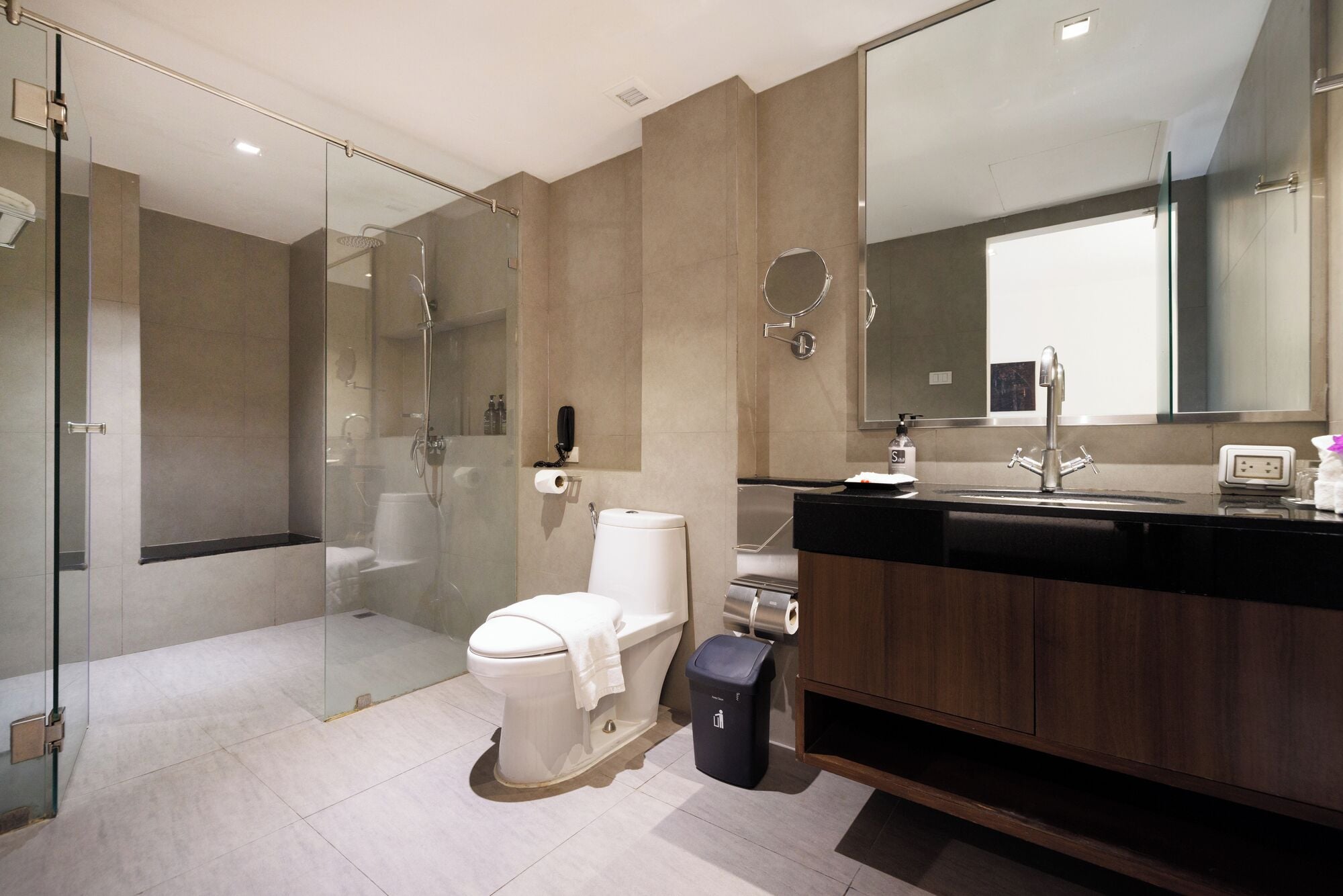 A modern bathroom with a glass-enclosed shower, a toilet, a sink with a mirror, and a trash bin.