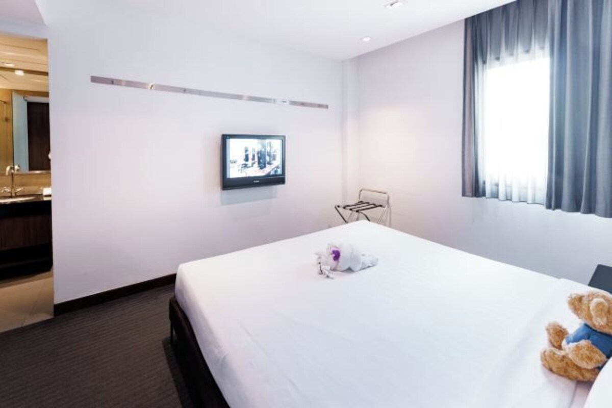 Una habitación de hotel con cama, televisión, ventana con cortinas y un osito de peluche sobre la cama.
