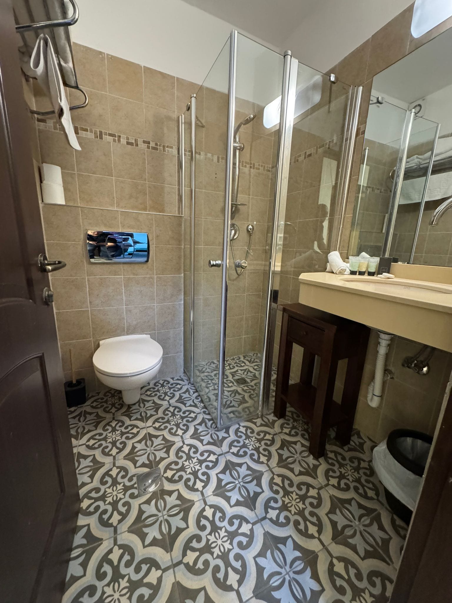 A bathroom with a glass-enclosed shower, a toilet, a sink with toiletries, and patterned floor tiles.