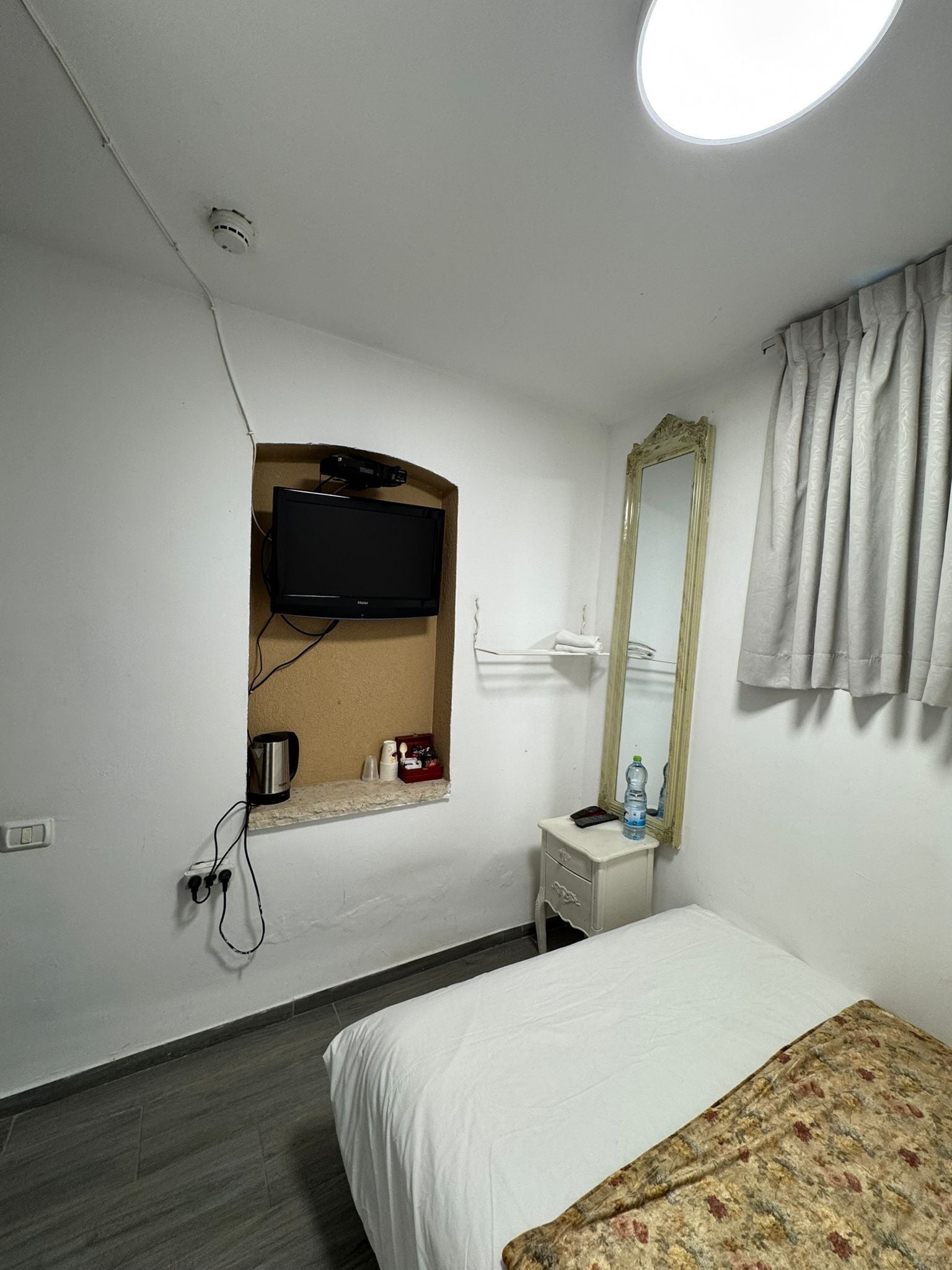 A hotel room with a bed, a television mounted on the wall, a mirror, a small table with a water bottle, and a power outlet.