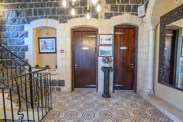 A stone-walled hallway with two wooden doors, a staircase with a metal railing, and a tiled floor.