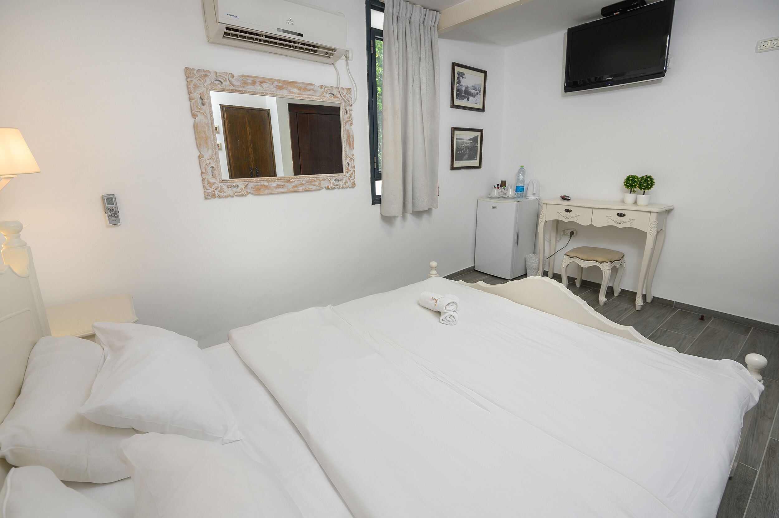 A neatly made bed with white linens, a bedside lamp, a small desk with a chair, a television mounted on the wall, and a framed mirror.