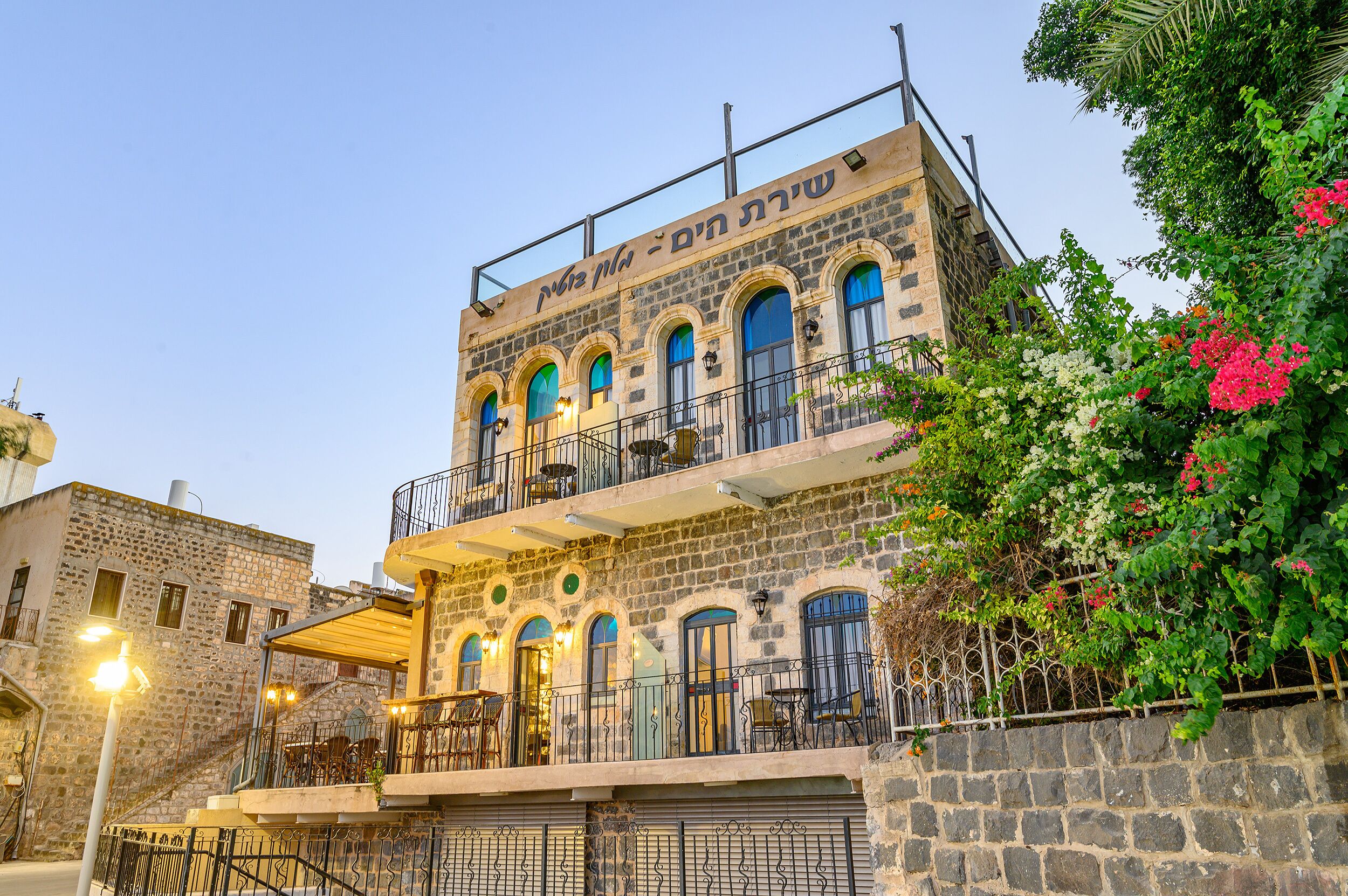 Un edificio de piedra con varios balcones y ventanas arqueadas, con mesas al aire libre y una escalera que conduce a la entrada.