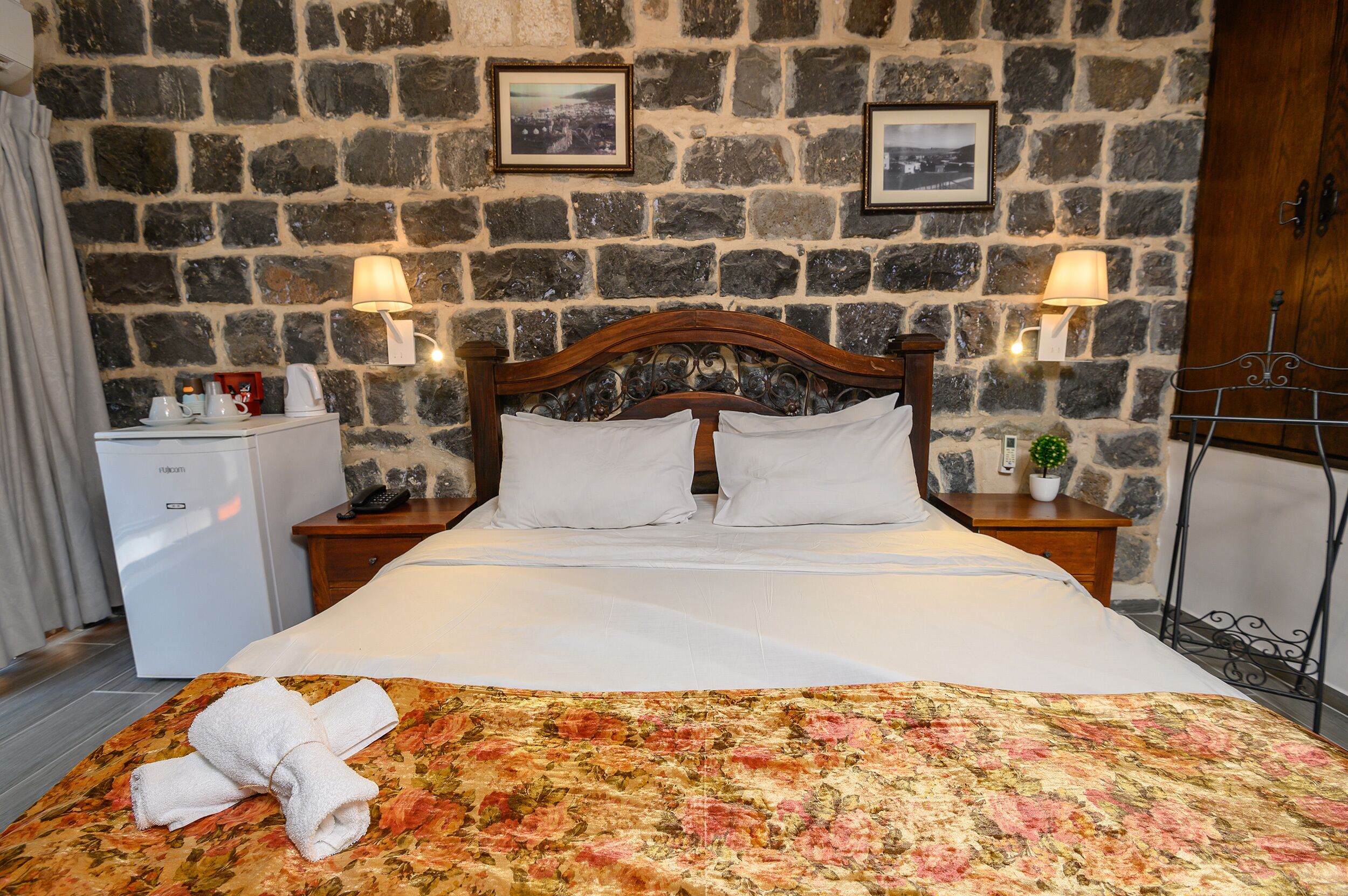 A neatly made bed with white linens, a floral-patterned bedspread, and a folded towel on the footboard. There are two bedside tables with lamps, a small refrigerator, and a wooden cabinet. The room features a stone wall with framed pictures.