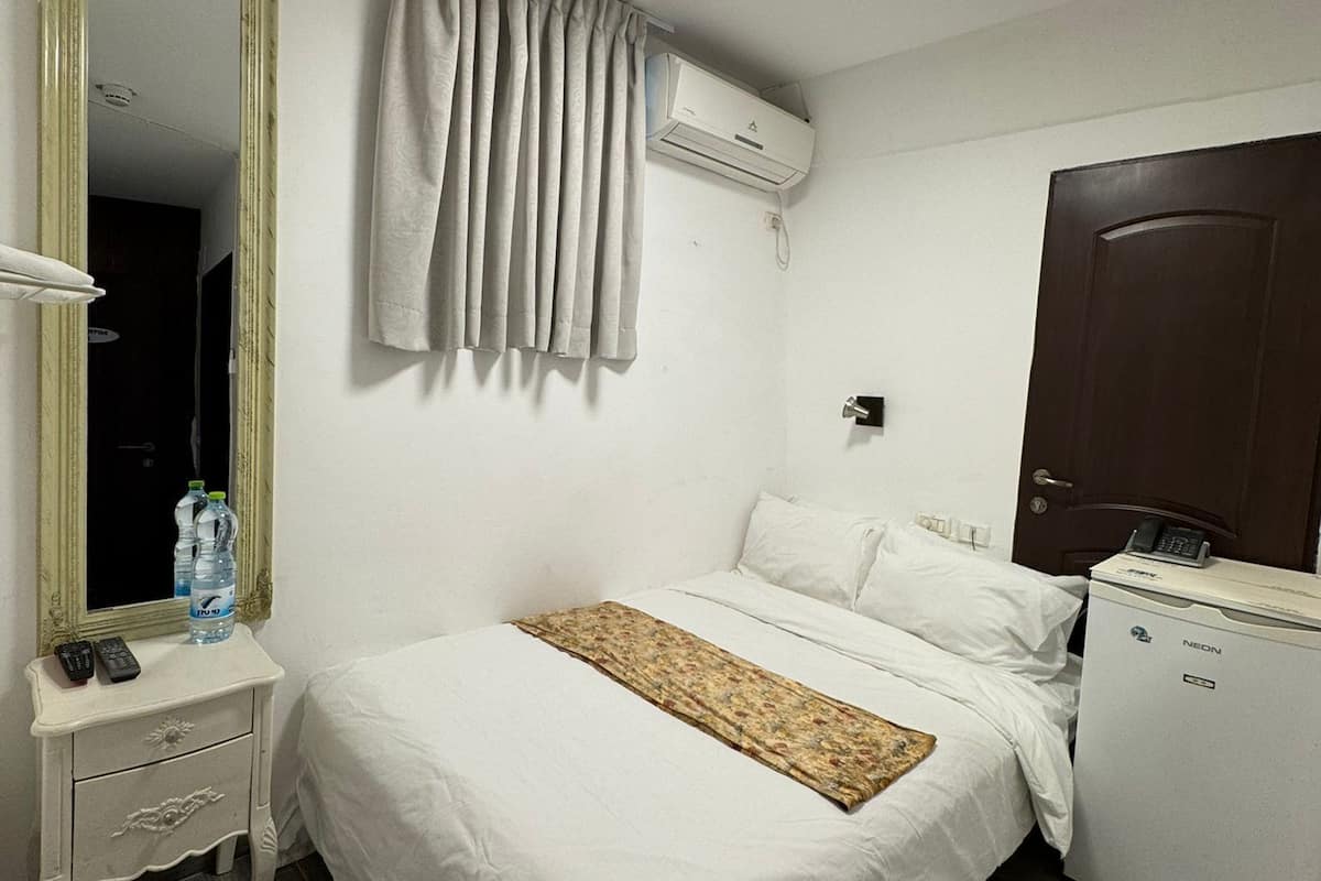 A hotel room with a bed, a bedside table with a remote control and water bottles, a mirror, a ceiling light, an air conditioning unit, and a small refrigerator.