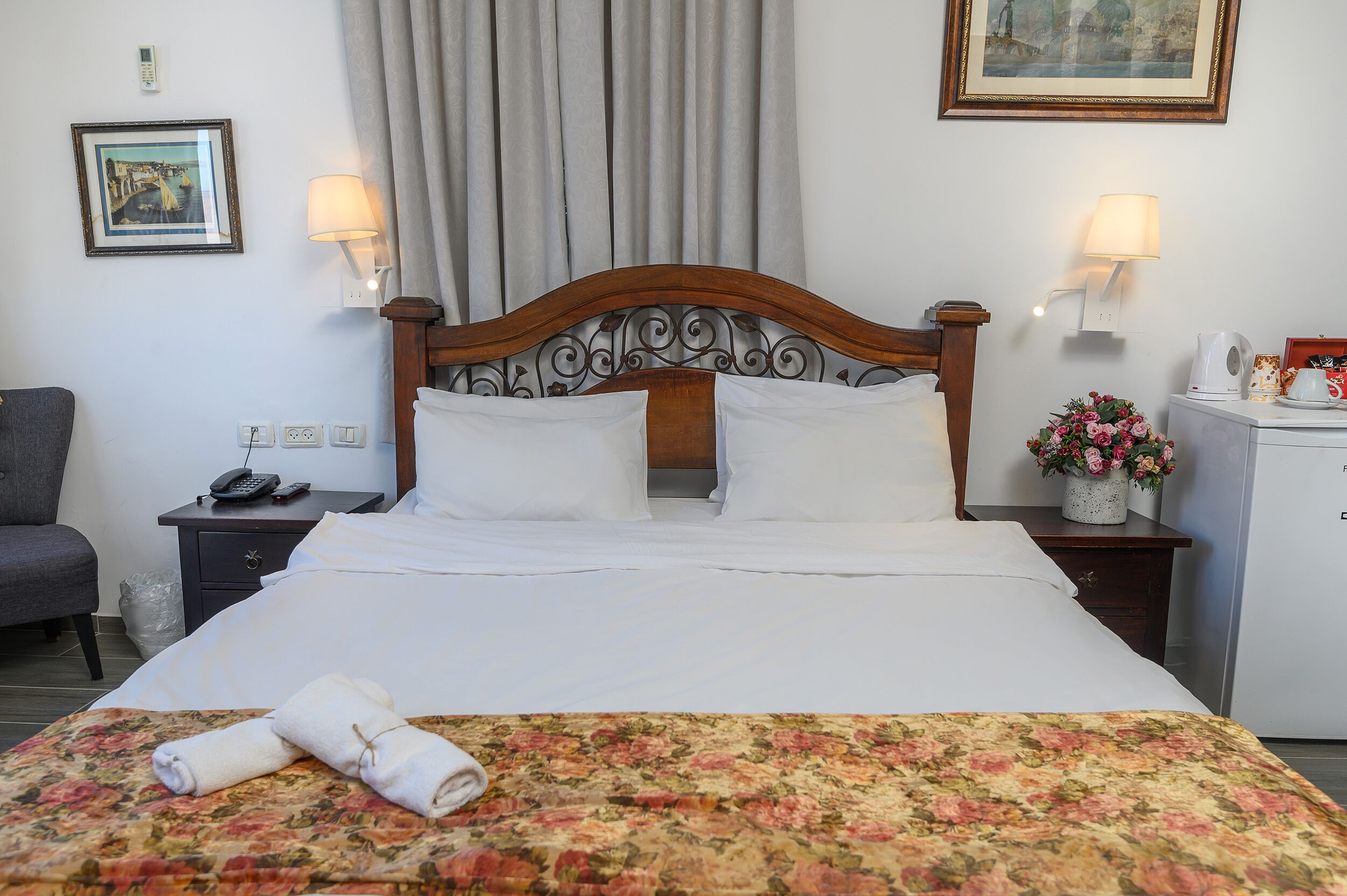 A neatly made bed with white linens, a floral bedspread, and a set of towels on the bed. There is a wooden headboard with a decorative design, two bedside lamps, and a small table with a vase of flowers. A telephone and a remote control are on the bedside table.