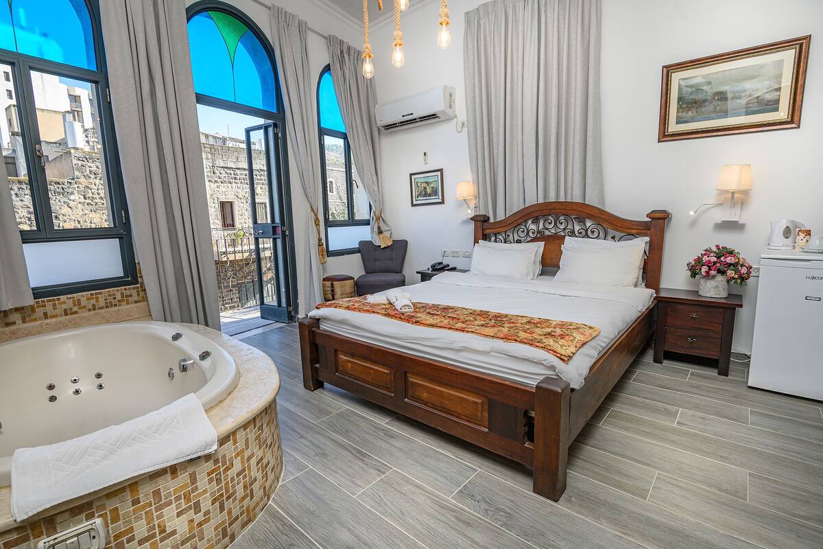 A hotel room with a large bed, a separate bathroom with a bathtub, a small desk, and a window with a view of buildings.