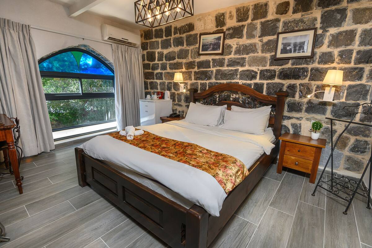A bedroom with a stone wall, a large bed, a nightstand, a lamp, and a window with curtains.