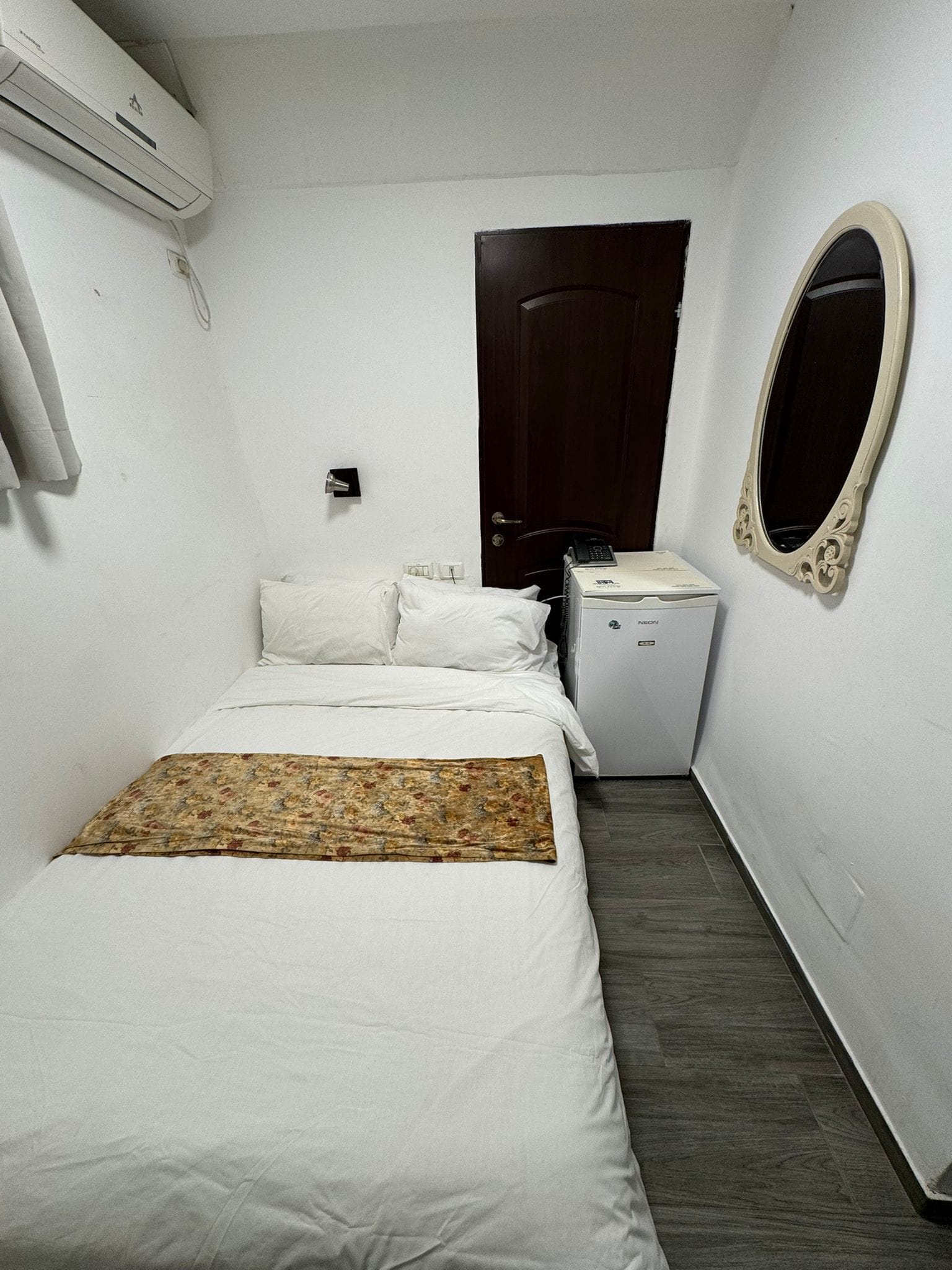 A small, clean hotel room with a bed, a bedside table, a mirror, and a small desk with a telephone.
