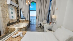 A hotel room with a large bed, a bathtub, a small table with a chair, and a view of the sea through a window. - Shirat Hayam - Boutique Hotel (Tiberias)