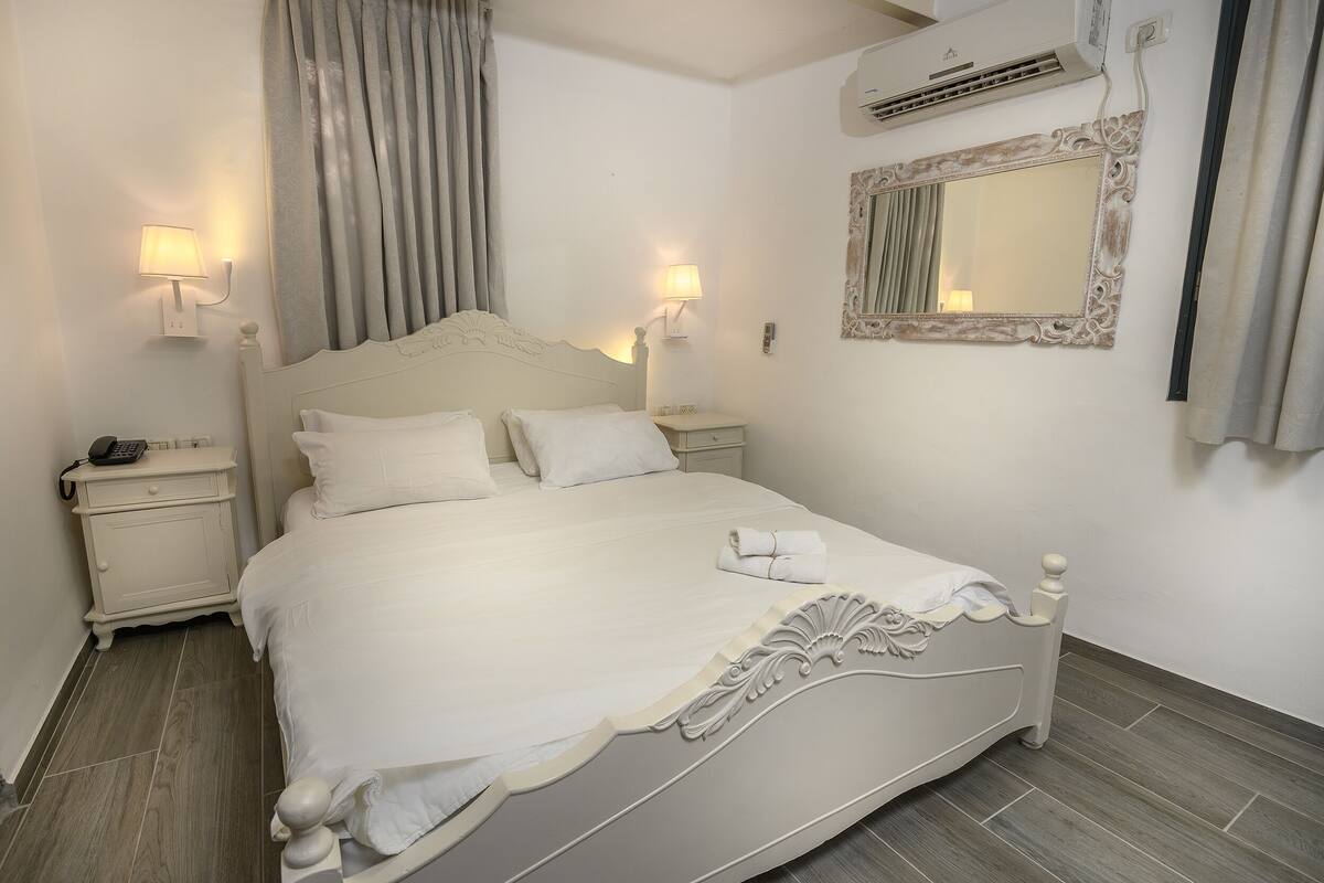 A neatly made bed with white linens, a bedside table with a lamp, and a wall-mounted mirror.