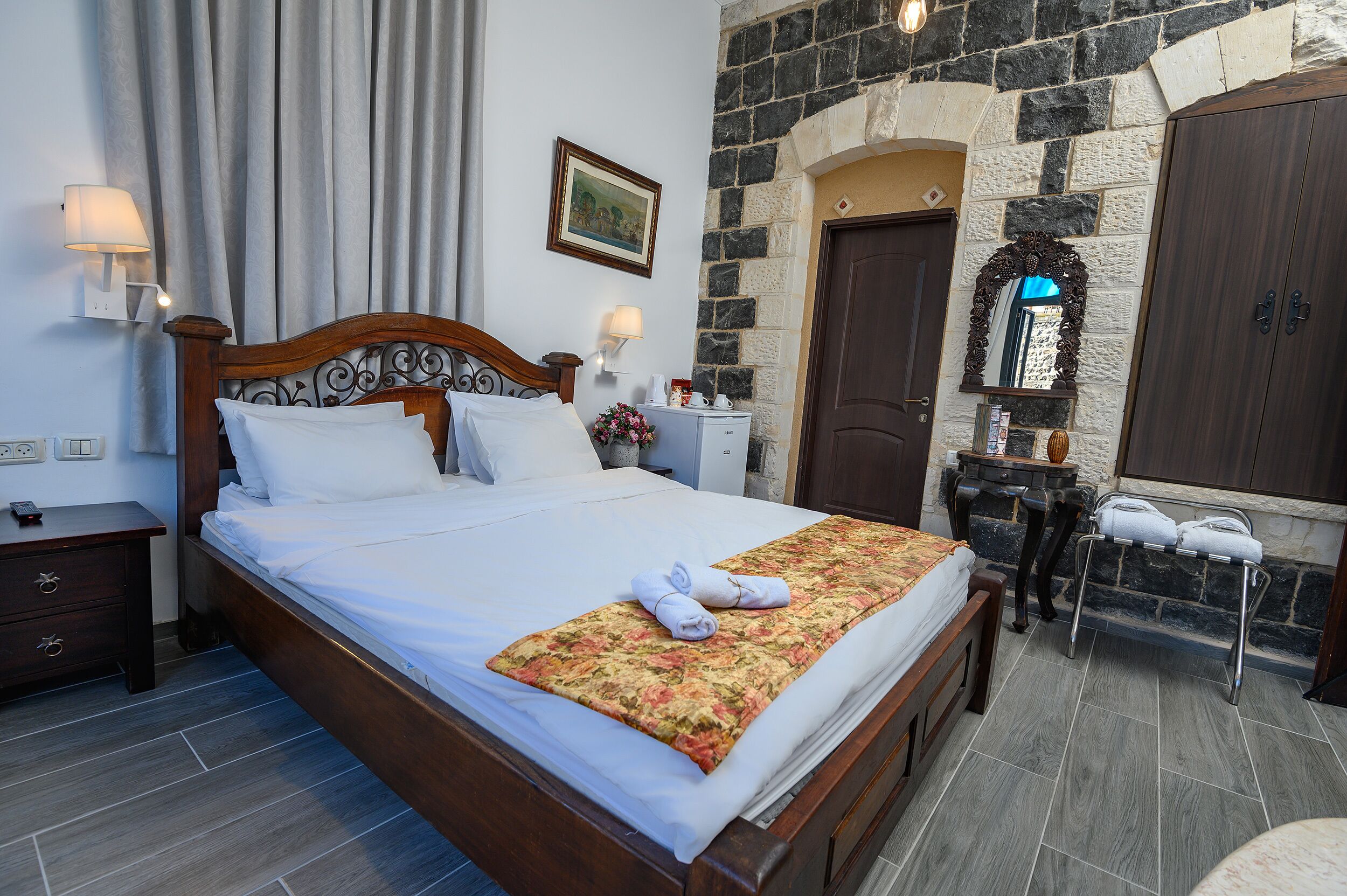 A neatly made bed with white linens, a wooden headboard, and a floral bedspread. There is a nightstand with a lamp and a small table with a mirror and a vase of flowers. The room has stone walls and a wooden door.