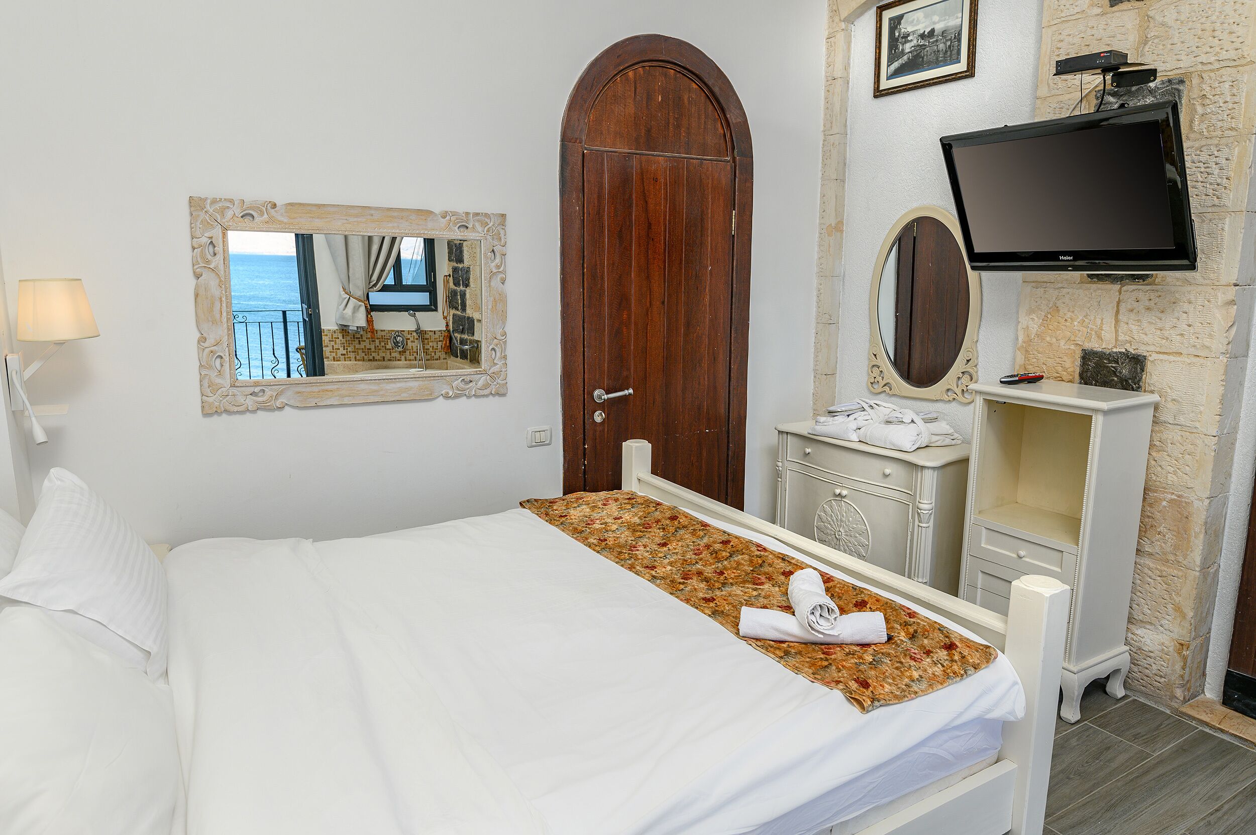 A hotel room with a bed, a wooden door, a television, a mirror, and a view of the sea.