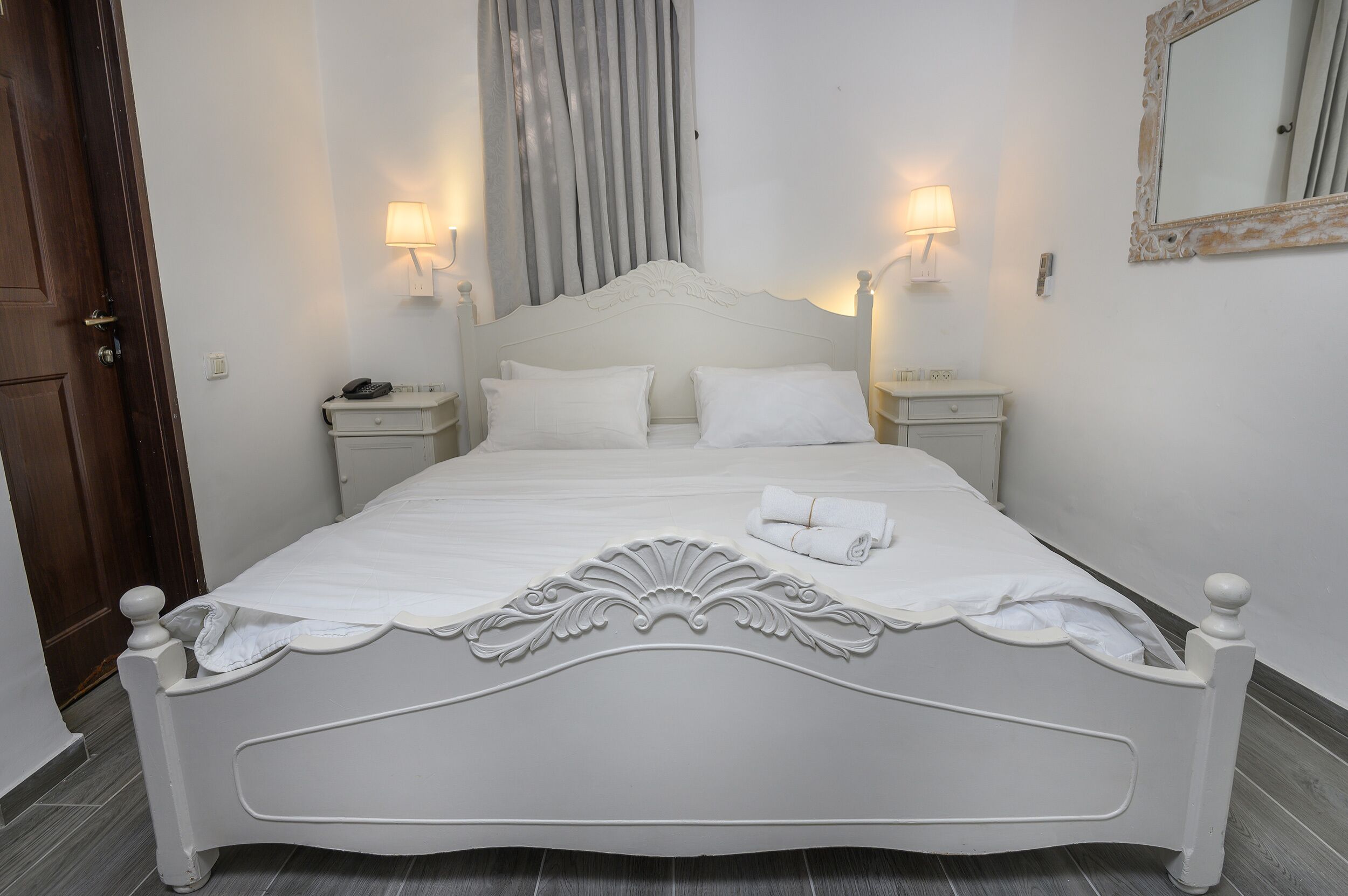 Una cama bien tendida con ropa blanca y un cabecero decorativo, dos lámparas de noche y un espejo en la pared.