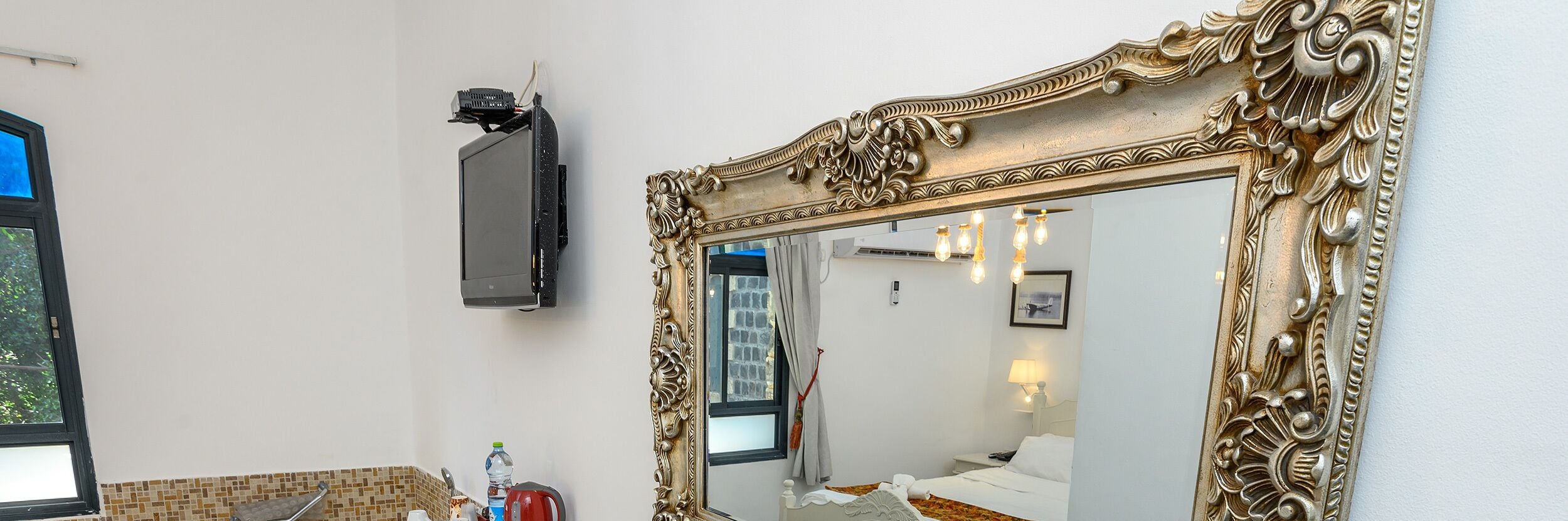A hotel room with a large ornate mirror, a flat-screen TV mounted on the wall, a bathroom with a tiled shower area, and a bed with white bedding.